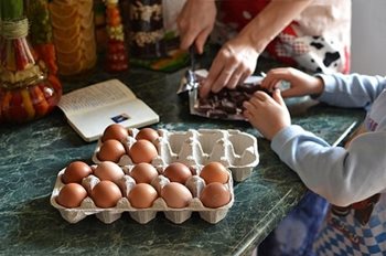 Adult and child hands working on a kitchen counter with eggs and a cookbook sitting out