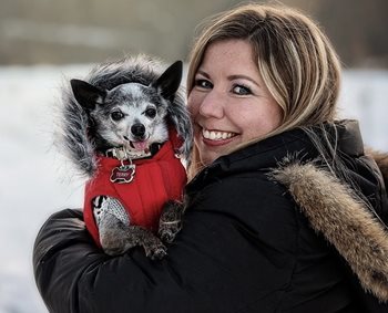 A woman smiling over her shoulder holding a small dog in a red coat on her shoulder