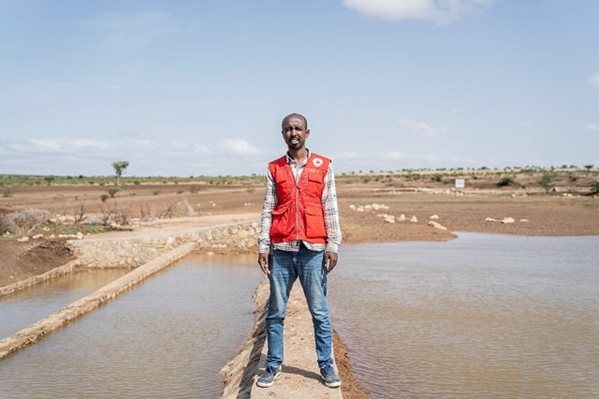 A man in a Red Cross standing in a field with a few reservoirs of water by his feet.