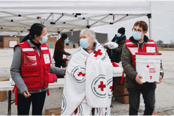 Two Red Cross members walking with an older woman wrapped in a Red Cross blanket
