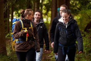 Two white men and two white women are wearing backpacks and warm jackets as they walk through a forest. They are smiling and talking together.