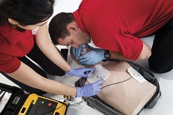 Two people in red shirts administering first aid techniques on a mannequin, including AED
