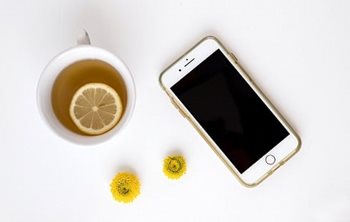 A phone beside a cup of tea with a slice of lemon floating in it.