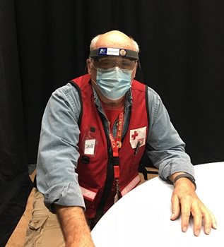 A man sitting at a table in a Red Cross vest and face shield