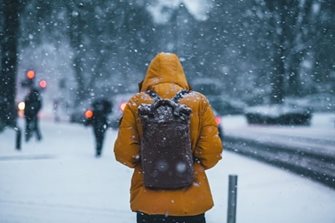 A person in a winter coat with hood up walking down a snowy sidewalk