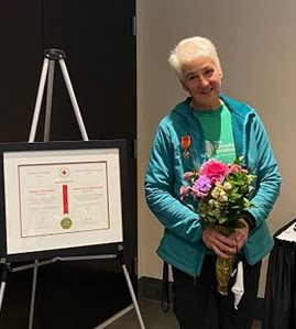 A woman with short white hair holding a bouquet of flowers standing beside a large framed award.