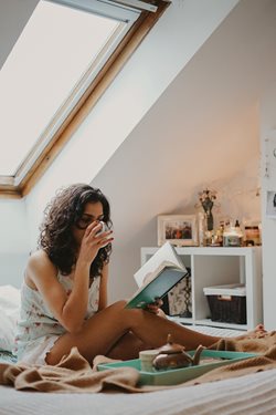 A woman is sitting on a bed, a cup of tea in one hand and a book in the other.