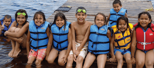 Nine children, ages approximately 7 - 11, wear life vests and bathing suits, smiling for a photo outdoors while sitting on a dock, on a lake.