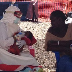 Woman in Ebola protective suit holds and feeds a one-month-old baby with Ebola as she sits beside the mother.