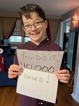 A young boy smiling while holding a handwritten note saying You did it! $10,000 raised!