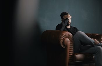 A man sitting down on a leather couch is holding his head with his hand, looking sad