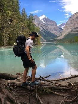 A white man in a white shirt, black shorts, blue hiking boots and a black backpack stands on the shore of a mountain lake in the Canadian Rockies