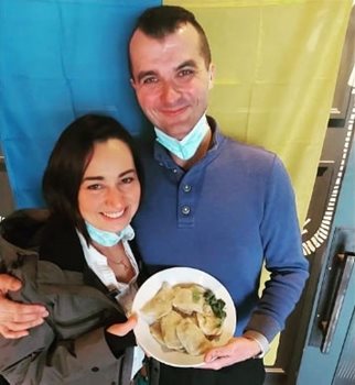 A man and woman standing in front of yellow and blue background holding a plate of pierogies