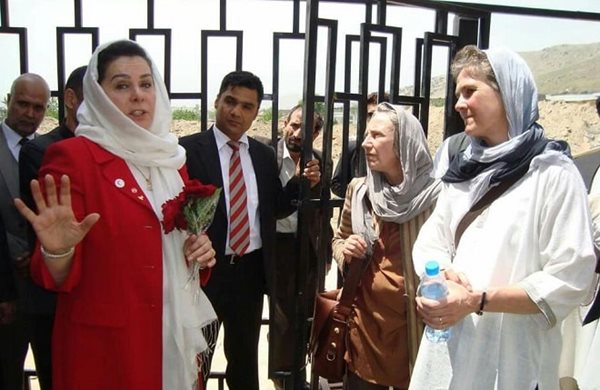 Pat, in a scarf to the right, standing among a crowd of people, listening to a woman in a scarf and red suit