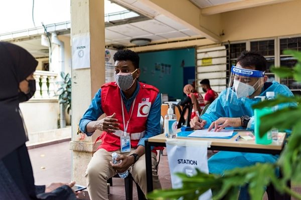 A woman in a mask sitting at a table talking to a man in mask in Red Cross vest.